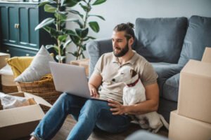 Young men moving in new house. He is happy and using laptop to organize everything. His pet dog is with him.
